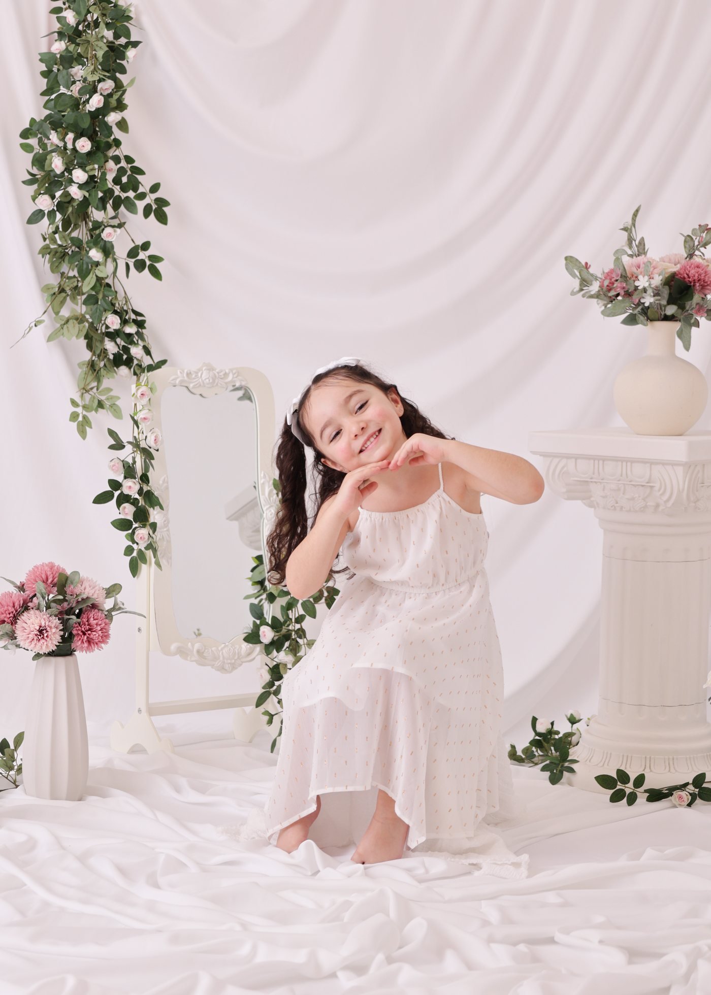 Little girl in a white dress among flowers and mirrors