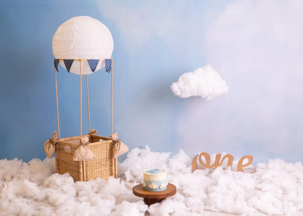 a setup or theme for baby photography with blue sky backdrop and a handmade hot air balloon and a "one" wooden prop and a wooden cake stand with a homemade beautiful cake with a little cloud hanged in the back and so much clouds on the floor or ground made for sitter babies and cake smash first birthday photoshoots